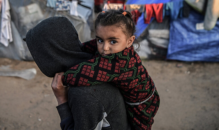 Kind in traditioneller Kleidung wird auf dem Rücken einer Person in Kapuzenjacke getragen, im Hintergrund Flüchtlingszelte in Gaza