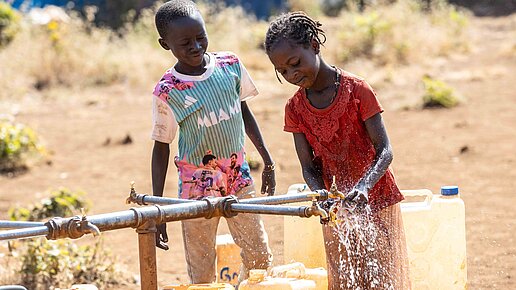Zwei Kinder füllen Kanister mit Wasser auf. Das Mädchen dreht den Hahn auf wodurch Wasser herausspritzt.