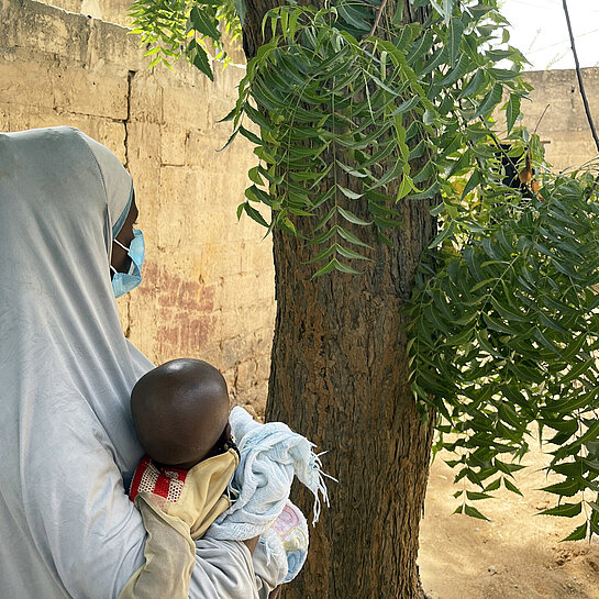 Eine junge Mutter mit langem Kopftuch hält ein Kind im Arm, sie ist seitlich von hinten fotografiert und nicht zu erkennen.