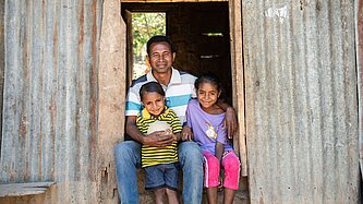 Ein Mann legt seine Arme um seine beiden Töchter, die mit ihm vor einer Hütte aus Wellblech sitzen