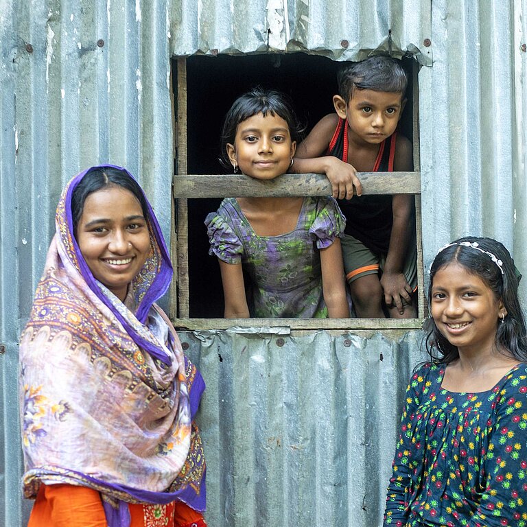 Zwei Mädchen in bunten Kleidern stehen vor einer Wellblechhütte, aus deren Fenster zwei kleine Kinder schauen