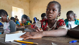 Ein Mädchen sitzt an einem Schulpult, auf dem ein Heft und Schreibmaterialien liegen.