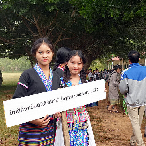 Nidsa heißt Besuch aus Japan an ihrer Schule willkommen Zwei junge Mädchen halten ein Schild mit laotischen Schriftzeichen. Sie stehen draußen.