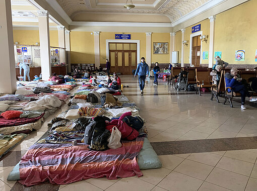 In einer Bahnhofshalle liegen Matratzen und Decken auf dem Boden, die als notdürftiger Schlafplatz für geflüchtete Menschen dienen.