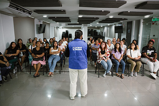 Eine Frau in einer blauen Weste von Plan International steht vor einer Gruppe von Frauen in einem Seminarraum. Alle sitzen und hören aufmerksam zu.