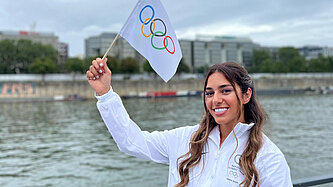 Die Sportlerin Saman Soltani steht am Ufer eines Flusses, trägt eine weiße Jacke mit dem Logo des Refugee Olympic Teams und hält eine Flagge mit den Olympischen Ringen in die Höhe. Sie lächelt in die Kamera.