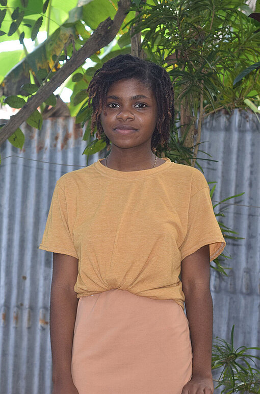 Stéphanie aus Haiti sorgt sich um die Bildungschancen von Mädchen in Haiti. Stéphanie steht vor einem Zaun und schaut lächelnd in die Kamera. Sie trägt ein apricotfarbenes Shirt und einen dazu passenden Rock.