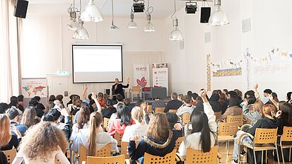Diana Dzaviza steht in einem Klassenraum und hält eine Präsentation. Die Schüler:innen hören gespannt zu.