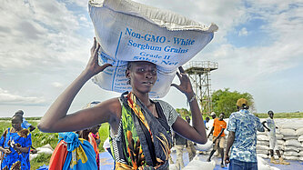 Ein Frau in buntem Kleid stemmt einen Sack Getreide auf ihren Kopf