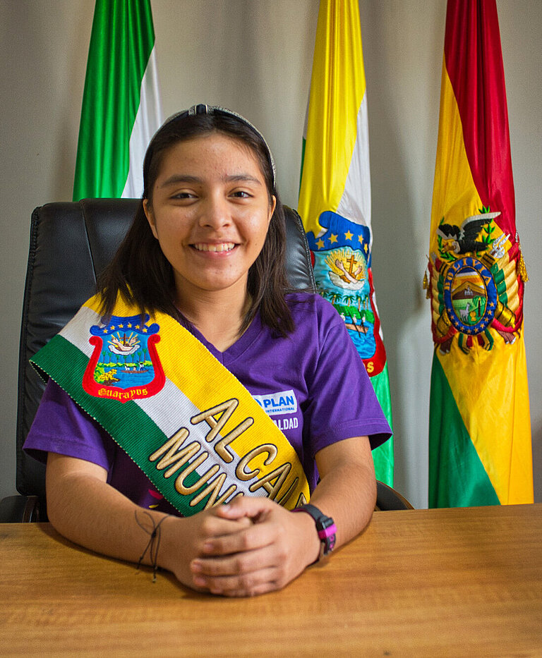 Ein Mädchen träg eine Schärpe und sitzt mit gefalteten Händen an einem Schreibtisch, hinter ihr sind drei Flaggen aufgestellt