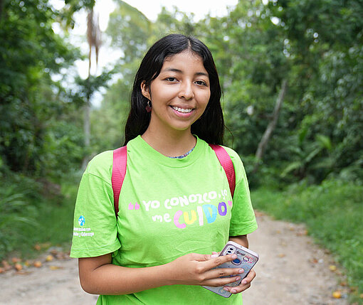 Eine junge Frau mit grünem T-Shirt steht auf einem sandigen Weg und blickt in die Kamera