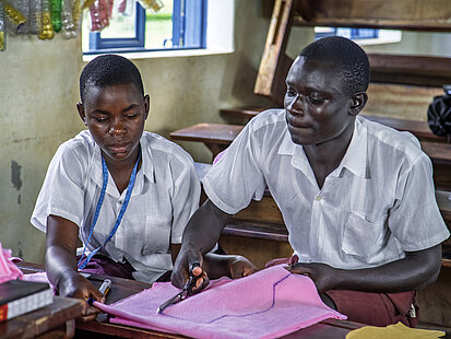 Zwei Jugendliche sitzen nebeneinander in einem Klassenraum, beide tragen weiße Hemden. Sie sind konzentriert und arbeiten gemeinsam an einem Bastelprojekt mit rosa Stoff, einer von ihnen schneidet mit einer Schere. 