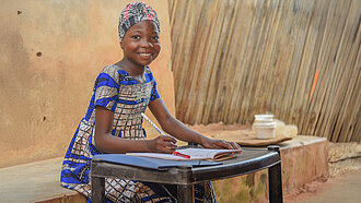 Ein Mädchen mit traditioneller, bunter Kleidung sitzt an einem kleinen Tisch. Vor ihr liegt ein Heft und sie hält einen Stift in der Hand. Sie lächelt in die Kamera.
