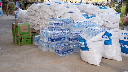 Gestapelte Kisten mit Speiseöl, Bündel von Wasserflaschen und große weiße Säcke mit Plan International-Logo liegen auf einer Betonfläche. Die Szene zeigt Hilfsgüter, die für die Verteilung vorbereitet sind.