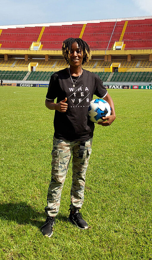 Tombody steht mit einem Fußball unter dem Arm in der Mitte eines Stadions und zeigt das Peace-Zeichen.