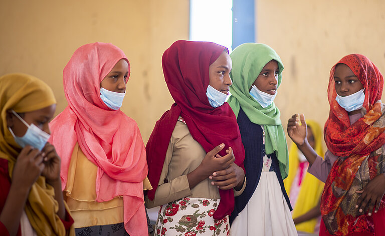Eine Gruppe Mädchen mit bunten Kleidern und Kopftüchern steht in einem Klassenzimmer.