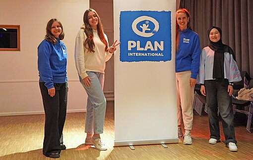 Drei junge Frauen stehen vor dem Logo der Kinderrechtsorganisation Plan International
